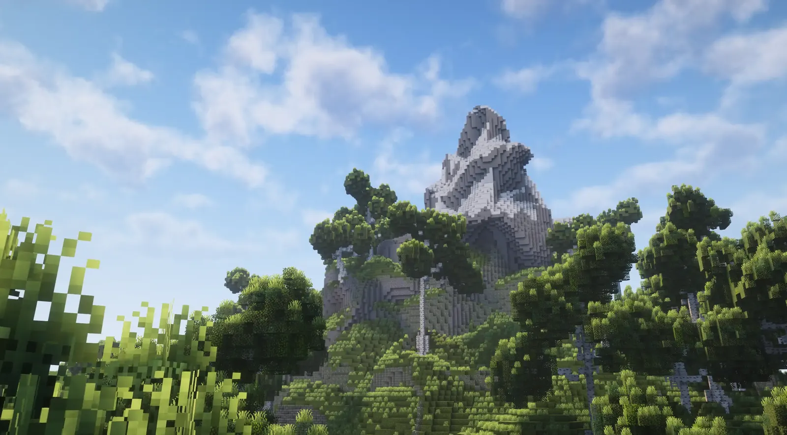 A rocky peak rising above a forest of blocky trees under a blue sky with clouds, in Minecraft with shaders.